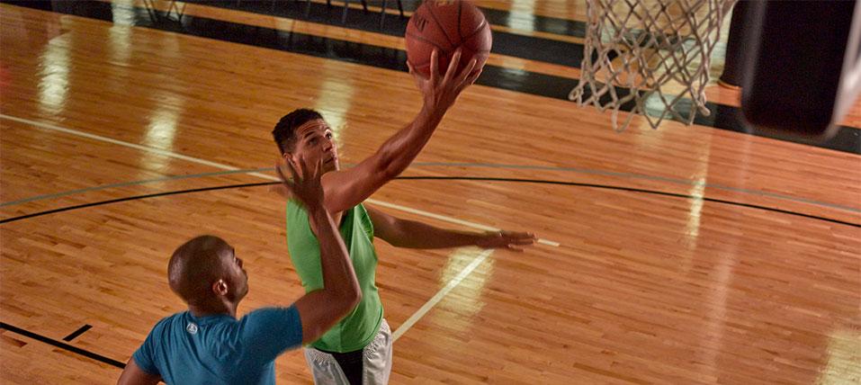 Two men playing basketball, one about to score against the other in a gym.