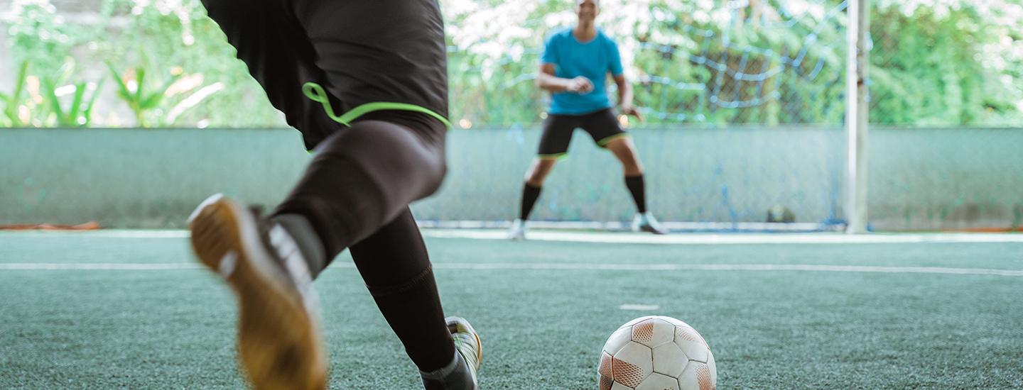 Player getting ready to kick a soccer ball with an opponent in front of a net in the background.