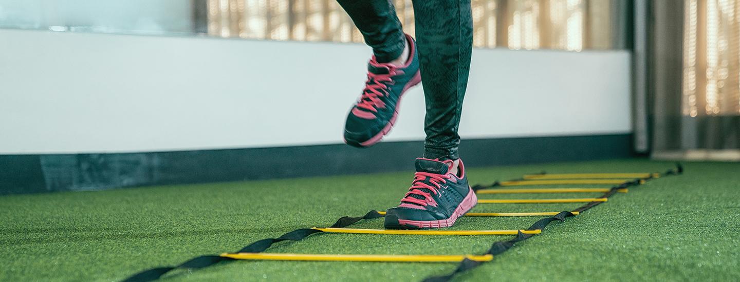 Woman performing agility training with a ladder on green turf in a gym, wearing black and pink athletic gear.