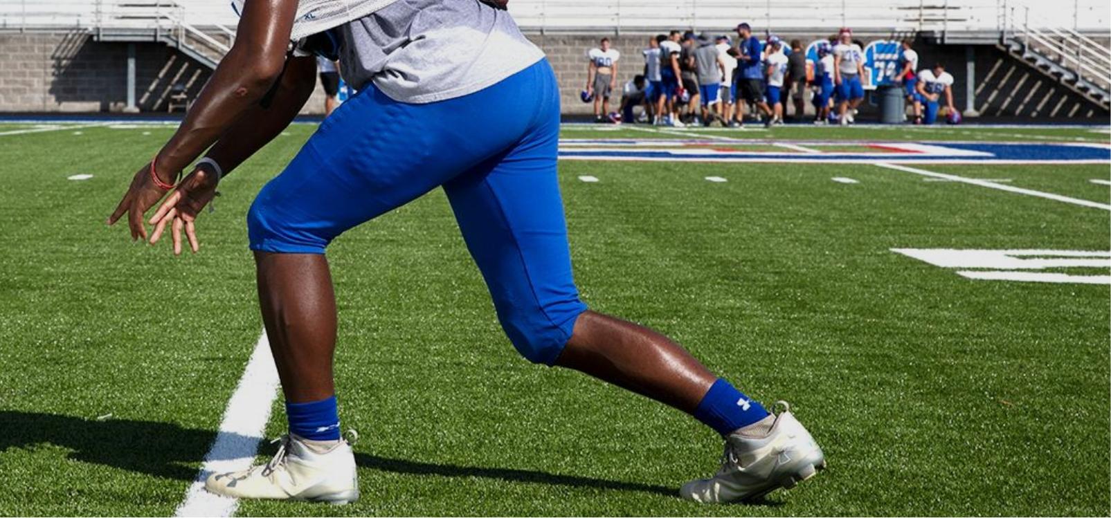 Football player in a stance on a field, preparing for a play, with teammates and opponents in the background.