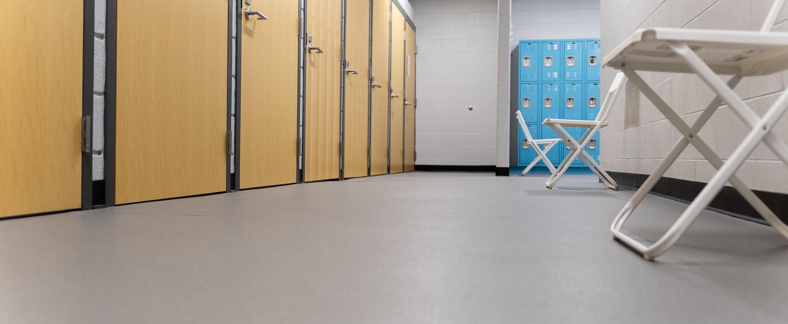 A shot of a locker room with changing rooms to the left, light blue lockers in the back and a white chair on the right