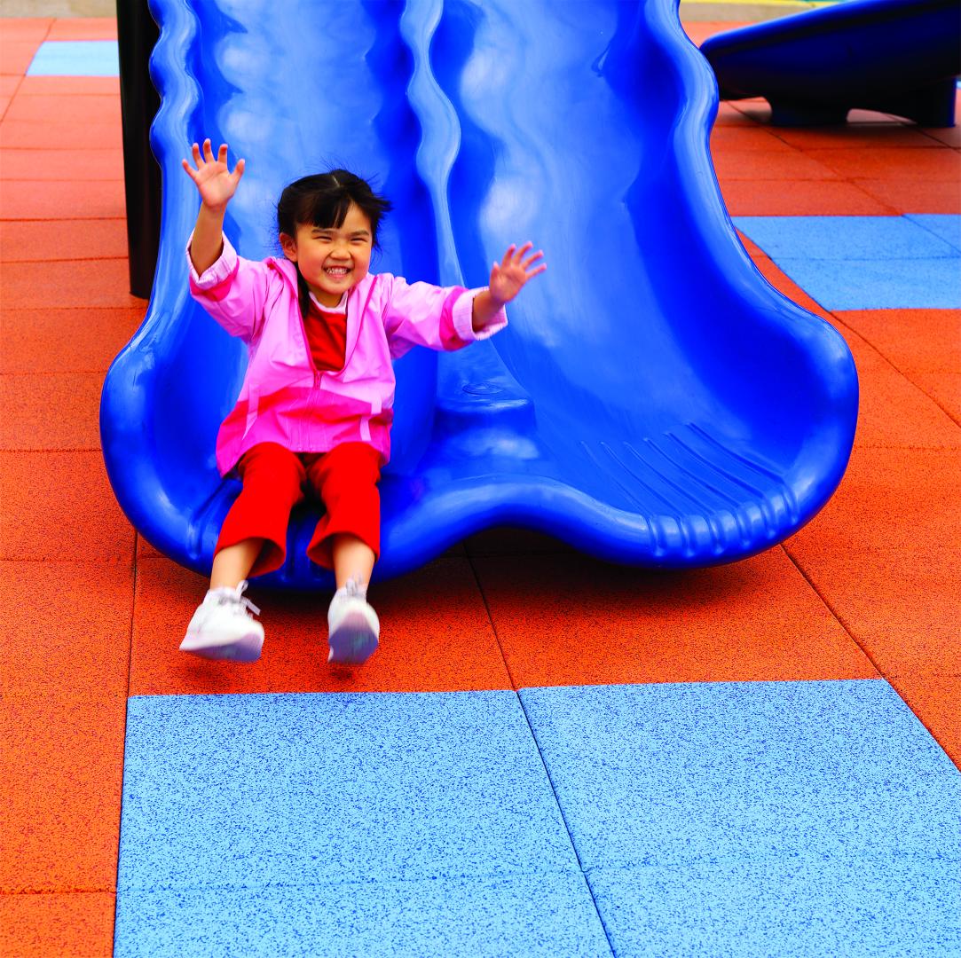 A young girl going down the slide very happy and landing on PlayGuard Tiles