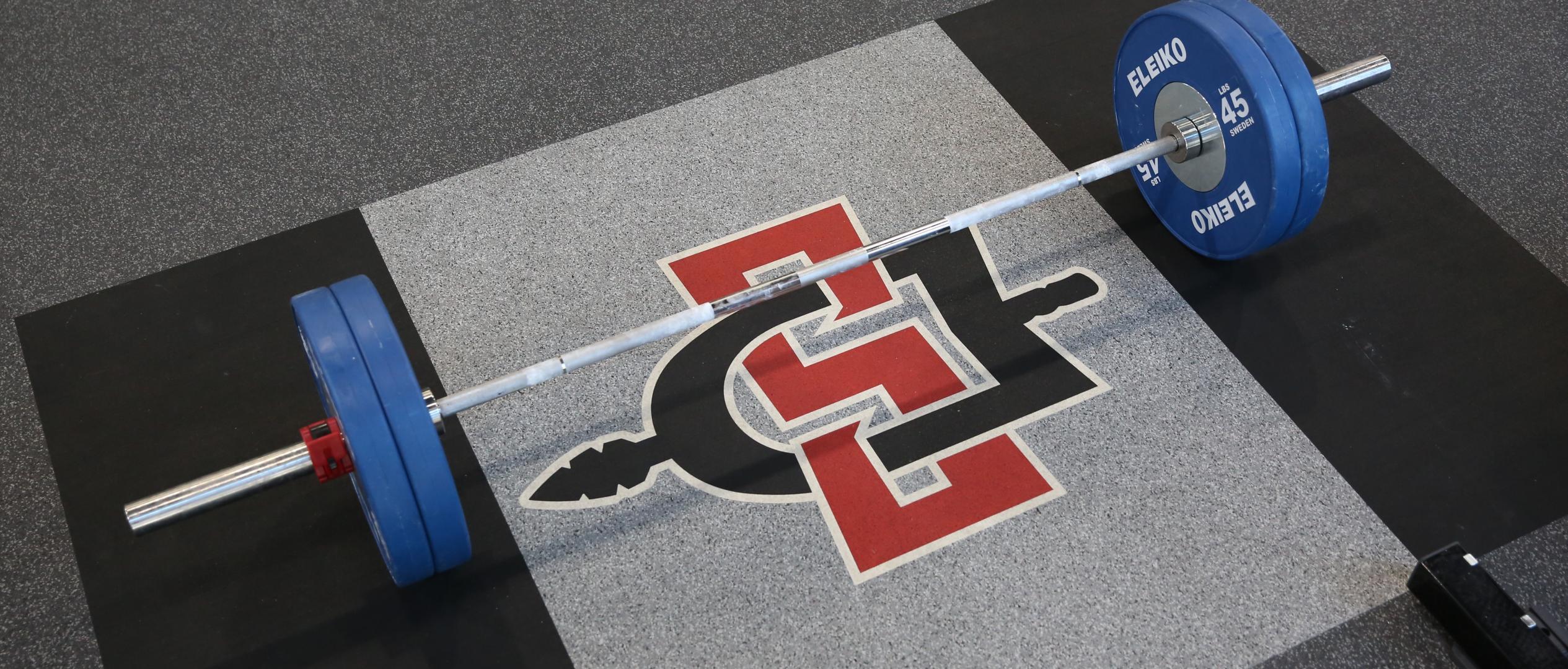 A lifting platform with a college logo in the middle and a barbell on top