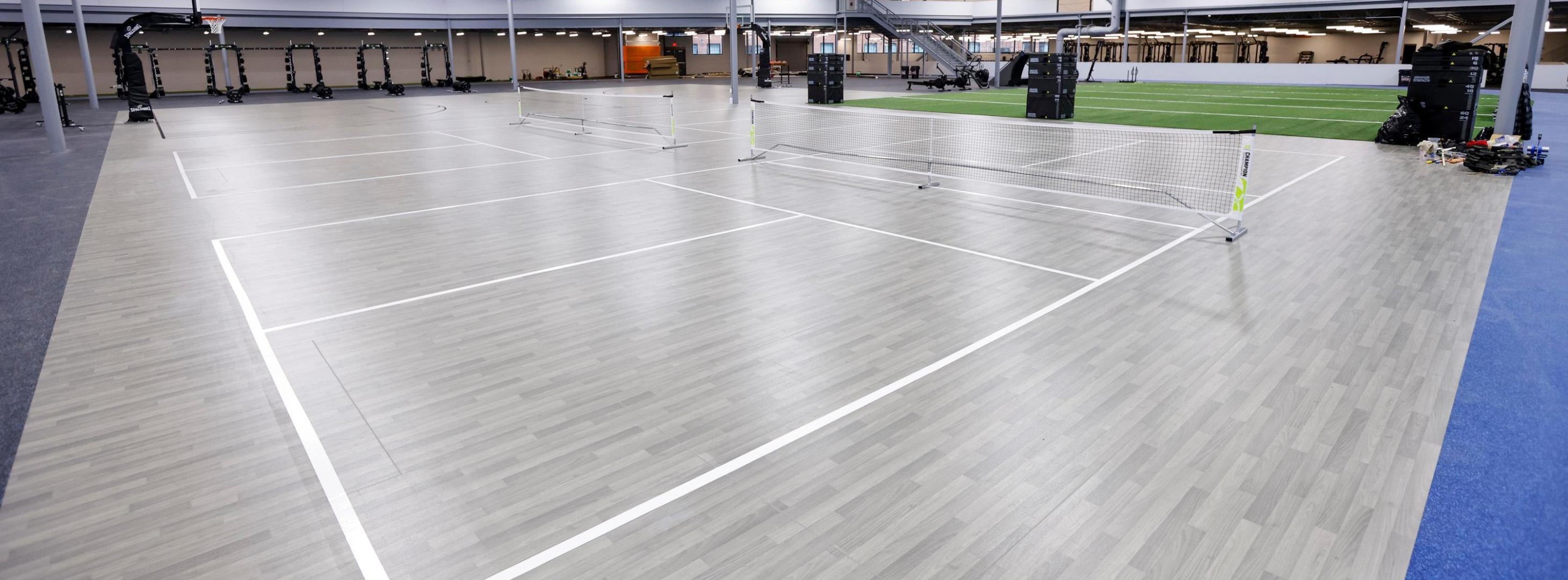 Two indoor courts side by side in an indoor fitness center with exercise equipment and artificial turf nearby. 